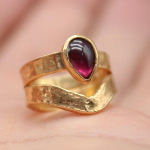 Op de afbeelding: Een gouden ring met een dieprode, druppelvormige edelsteen. De ring heeft een getextureerde band en een chevron-ontwerp. De ring wordt op een hand getoond. Het logo van Classi Fine Jewelry staat linksonder.