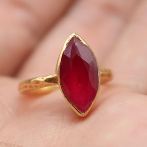 May include: A gold ring featuring a marquise-cut, deep red gemstone. The ring is held in a hand, highlighting the detailed setting and the vibrant colour of the stone. The ring is from Classi finejewelry.