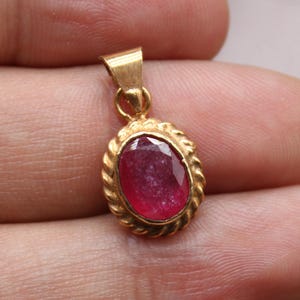 May include: A gold pendant featuring a deep red, oval gemstone. The pendant has a twisted rope-style border and a small loop for a chain. The pendant is held in a hand, with the "Classi finejewelry" logo in the lower left corner.