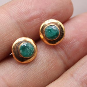 Puede incluir: Un par de pendientes de botón dorados, cada uno con una piedra preciosa verde redonda y vibrante engastada en un bisel dorado. Los pendientes son pequeños y delicados. Los pendientes se muestran en una mano.