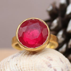 May include: A gold ring with a large, round, faceted red gemstone. The ring is displayed on a seashell, with a pine cone in the background. The logo "Classi finejewelry" is in the lower left corner.