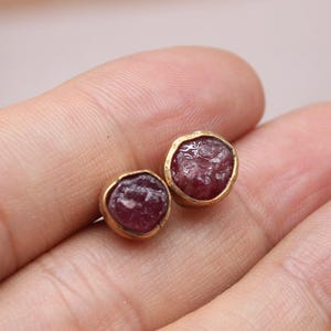 Op de afbeelding: Een paar goudkleurige oorstekers met ronde, ruw geslepen, rood-paarse edelstenen. De oorbellen worden in een hand gehouden, met een vage achtergrond van houten blokken en een logo in de linkerbenedenhoek.