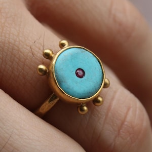 Op de afbeelding: Een gouden ring met een ronde turquoise steen en een kleine rode steen in het midden. De ring heeft een decoratief ontwerp rond de turquoise steen. De ring zit om een vinger. Het logo van Classi finejewelry staat linksonder.