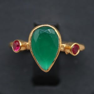 May include: A gold ring featuring a large, pear-shaped green gemstone as the centerpiece, flanked by two smaller, round red gemstones. The ring is displayed on a black surface, showcasing the jewelry's design and color contrast. The ring is a piece of fine jewelry.