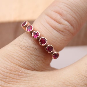 May include: A rose gold ring set with seven round, vibrant red gemstones. The ring is displayed on a person's finger, showcasing the jewelry's design and color. The ring is from Classi Fine Jewelry.