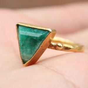 May include: A gold ring with a triangular, emerald-green gemstone. The ring is held in a hand, with the focus on the jewelry. The ring has a gold band and setting. The logo for Classi finejewelry is in the lower left corner.