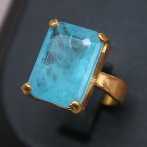 Pode incluir: Um anel dourado com uma grande pedra preciosa retangular azul clara. A pedra é engastada numa moldura dourada com quatro garras de canto. O anel é exibido numa superfície preta. A marca "Classi finejewelry" é visível.