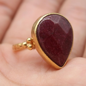 May include: A gold ring with a large, teardrop-shaped, dark red gemstone. The ring has a textured band with small gold beads. The gemstone is set in a gold bezel. The ring is displayed on a hand.