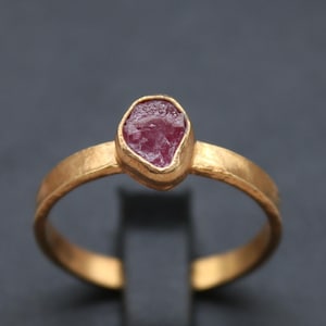 May include: A gold ring with a raw, pink gemstone set in a bezel. The ring has a textured band and is displayed on a black stand. The jewelry is from Classi finejewelry.