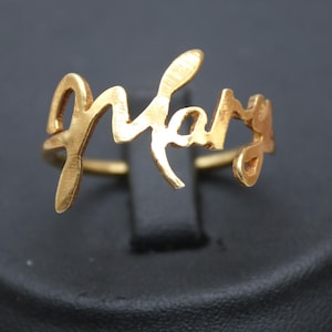 May include: A gold-colored ring spelling out the name "Mary" in a cursive font. The ring is displayed on a black surface, with the "Classi finejewelry" logo visible in the lower left corner.