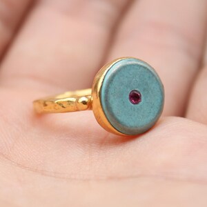 May include: A gold ring with a round, teal-colored stone and a small red accent in the center. The ring is held in a hand, showcasing the intricate details of the jewelry. The ring's band has a textured design.