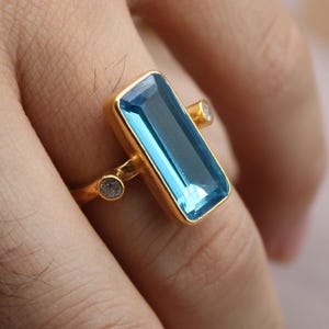 May include: A gold ring featuring a rectangular, light blue gemstone as the centerpiece. Two smaller, round gemstones flank the main stone, with a gold band. The ring is displayed on a hand, showcasing the jewelry's design.