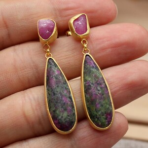 May include: Gold-toned earrings with a teardrop-shaped, speckled green and purple stone pendant. The earrings also feature a small, rectangular pink stone at the top. The earrings are held in a person's hand. The logo for Classi finejewelry is in the bottom left corner.