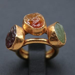 May include: A gold ring featuring three raw gemstones in shades of red, orange, and green. The ring has a double-band design and is displayed on a black stand. The ring is from Classi Fine Jewelry.
