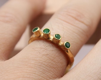 Puede incluir: Un anillo de oro con cuatro piedras preciosas verdes redondas. El anillo se lleva en un dedo. El anillo es una joya. El anillo es de color oro.