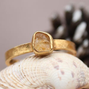 Op de afbeelding: Een goudkleurige ring met een rechthoekige, lichtgele edelsteen. De ring ligt op een zeeschelp, met een vage achtergrond met een dennenappel en een houten oppervlak. De ringband heeft een getextureerde afwerking.