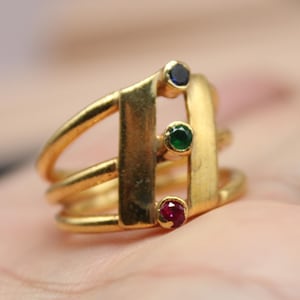 May include: A gold-colored ring with three bands and three colored gemstones: a dark blue, a green, and a red. The ring has a unique design with vertical bars and is displayed on a hand. The logo "Classi finejewelry" is in the lower left corner.