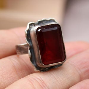 Puede incluir: Un anillo de plata con una piedra preciosa rectangular de color rojo intenso. La piedra preciosa está engastada en un bisel texturizado y adornado. El anillo se muestra en una mano, mostrando el diseño y el color de la joya.