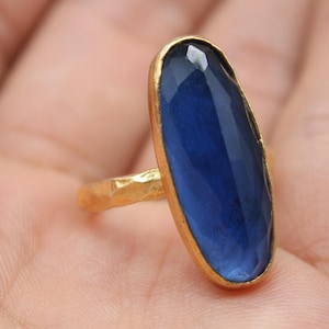 Peut inclure: Une bague dorée ornée d'une grande pierre précieuse ovale bleu foncé. La bague a un anneau texturé et un sertissage en or. L'image comprend le logo "Classi finejewelry" dans un carré noir.