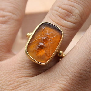 May include: A gold ring with an amber-colored, rectangular stone featuring an insect carving. The ring is on a person's finger. The ring has a gold band and small gold accents on the sides. The ring is a piece of fine jewelry.