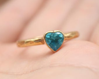 May include: A gold ring with a heart-shaped, teal-colored gemstone. The ring is held in a person's hand, with the focus on the jewelry. The ring has a hammered texture. The logo "Classi finejewelry" is in the lower left corner.