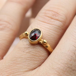 May include: A gold ring with a dark red oval gemstone. The ring has a simple band with small decorative beads on either side of the gemstone. The ring is on a finger.