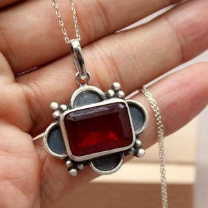 May include: A silver pendant necklace featuring a rectangular, deep red gemstone. The pendant is set in a decorative silver frame with a dark blue background. The necklace is held in a hand.