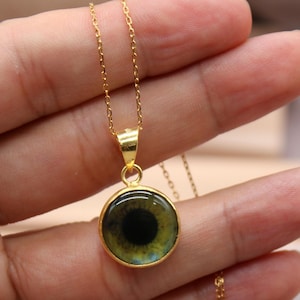 May include: Gold-colored necklace with a pendant featuring a detailed eye design. The pendant is round, with a black pupil and green iris, set in a gold frame. The chain is delicate and also gold-colored.