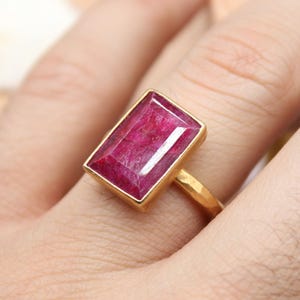 May include: A gold ring featuring a large, rectangular, deep pink gemstone. The ring has a simple, elegant design with a hammered gold band. The gemstone is set in a gold bezel, creating a striking contrast. The ring is a piece of fine jewelry.