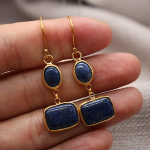 May include: Gold-toned earrings with a drop design, featuring oval and rectangular dark blue gemstones. The earrings are held up against a neutral background, showcasing the jewelry's color and shape.