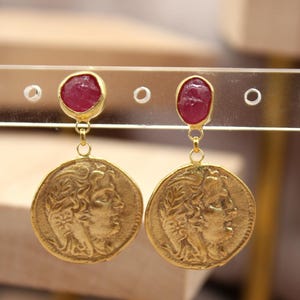 Pode incluir: Brincos dourados com um design semelhante a uma moeda e uma pedra preciosa vermelha no topo. Os brincos apresentam um perfil de uma pessoa na moeda. Os brincos são exibidos em um suporte transparente.