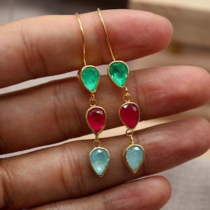 Peut inclure: Boucles d'oreilles pendantes dorées avec trois pierres précieuses en forme de goutte, vertes, rouges et bleu clair. Les boucles d'oreilles sont présentées par une main. Le logo "Classi finejewelry" est visible.