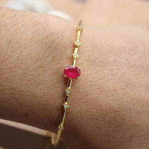 May include: A gold bracelet with a central oval ruby and small diamond accents. The bracelet is worn on a person's wrist, showcasing the fine jewelry's design and craftsmanship. The bracelet's design is delicate and elegant.