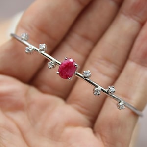 Puede incluir: Una pulsera de plata con una gema de rubí ovalada y pequeñas gemas claras. La pulsera tiene un diseño lineal delicado con las gemas engarzadas a lo largo de la banda. El rubí es de color rojo vibrante.