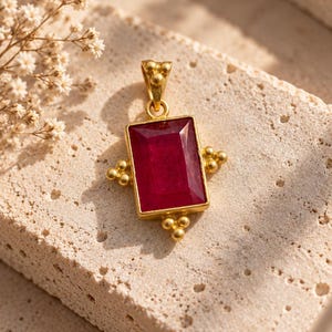 May include: A rectangular pendant featuring a deep red gemstone set in a gold-toned metal frame. The setting includes small decorative gold spheres on the sides and bottom, with a matching gold bail at the top for attaching to a chain or necklace.