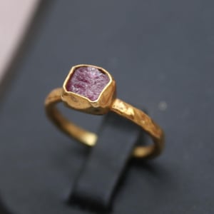 Puede incluir: Un anillo de oro con una piedra preciosa en bruto, sin tallar, de color rojo rosado intenso, engastada en un bisel de oro martillado y texturizado. La banda del anillo también muestra una textura de superficie martillada e irregular.