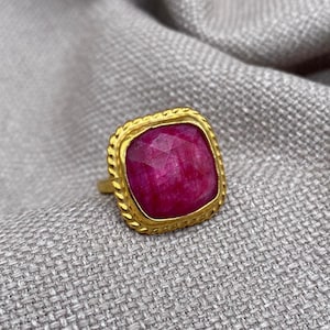 Puede incluir: Un anillo de oro con una gran piedra preciosa de color rosa intenso, tallada en forma cuadrada y facetada. El engaste tiene un borde texturizado similar a una cuerda que rodea la piedra, la cual está montada en un marco de oro pulido.