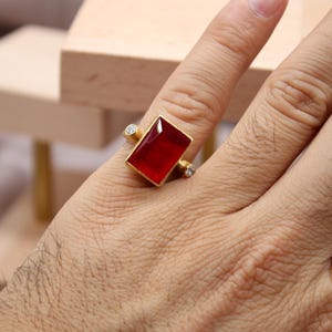 May include: A gold ring featuring a large rectangular red gemstone set in the center, flanked by two small round clear stones on either side. The band has a matte gold finish, and the design is presented on a finger with a logo in the corner.