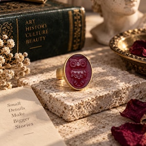 Peut inclure: Une bague dorée ornée d'une pierre ovale rouge foncé translucide avec un motif de hibou en relief. La bague présente un anneau en métal lisse et poli ainsi qu'un sertissage clos qui maintient la pierre centrale aux détails sculptés.