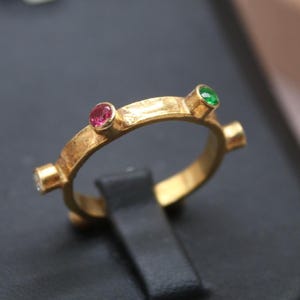 May include: A gold ring with a textured, hammered finish featuring multiple bezel-set round gemstones. The visible stones include a vibrant pink gem and a bright green gem, each secured in a raised setting along the circular band.