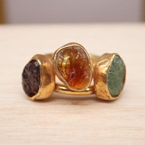 Puede incluir: Un anillo de oro con tres piedras preciosas en bruto engastadas y dispuestas horizontalmente. Las piedras incluyen una gema de color marrón oscuro a la izquierda, una gema de color ámbar en el centro y una gema de color verde claro a la derecha.