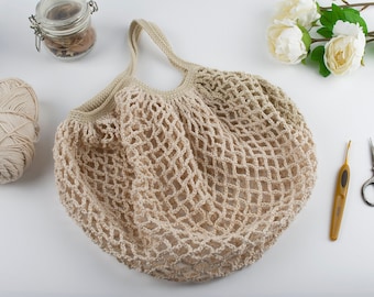 May include: A beige, crocheted net bag with two handles, perfect for carrying groceries or other items. The bag is made of a light-colored yarn and has a large, open weave. A ball of yarn, scissors, and a crochet hook are also visible.