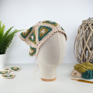 May include: A beige, green, and teal crochet headscarf with a geometric pattern. The head covering is displayed on a mannequin head. The headscarf has a scalloped edge and ties. Additional crochet pieces and yarn are visible.