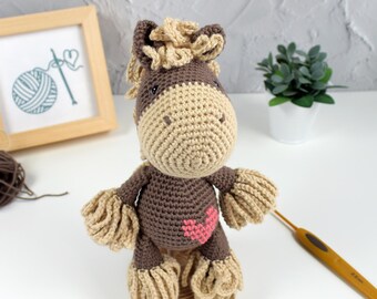 Op de afbeelding: Een bruin en beige gehaakt paard met een roze hart op zijn borst. Het paard staat op een houten basis en heeft een gehaakte manen en staart.
