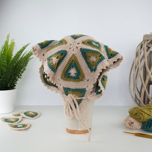 May include: A beige, green, and olive green crochet hat with a triangular pattern. The hat has a scalloped edge and a tie closure. Additional crochet pieces are displayed nearby. The hat is displayed on a mannequin head.