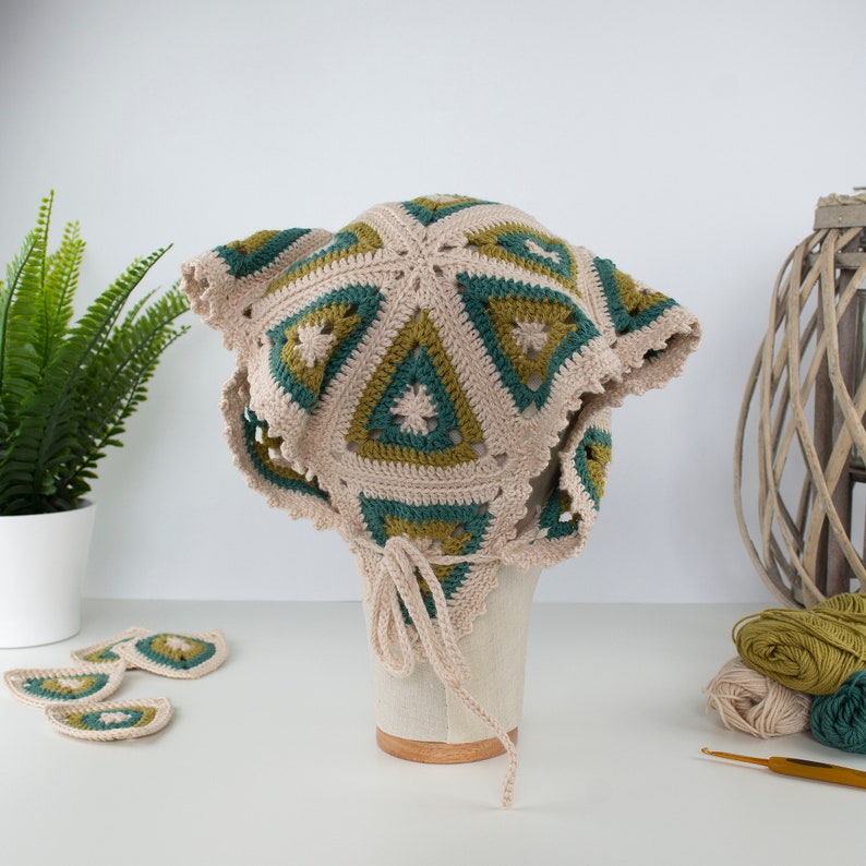 May include: A beige, green, and olive green crochet hat with a triangular pattern. The hat has a scalloped edge and a tie closure. Additional crochet pieces are displayed nearby. The hat is displayed on a mannequin head.