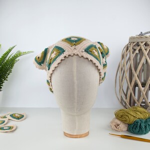 May include: A crocheted head covering in a square pattern, featuring cream, green, and olive green colors. The hat is displayed on a mannequin head. Crochet yarn and a crochet hook are visible in the background.