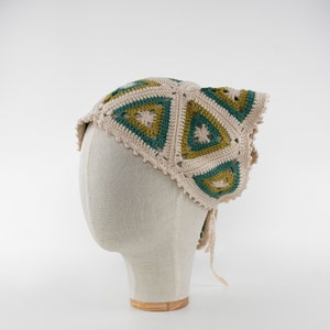 May include: A cream-colored crochet headscarf with a geometric pattern of green and olive triangles. The head covering is displayed on a mannequin head and has a scalloped edge and tie closures.