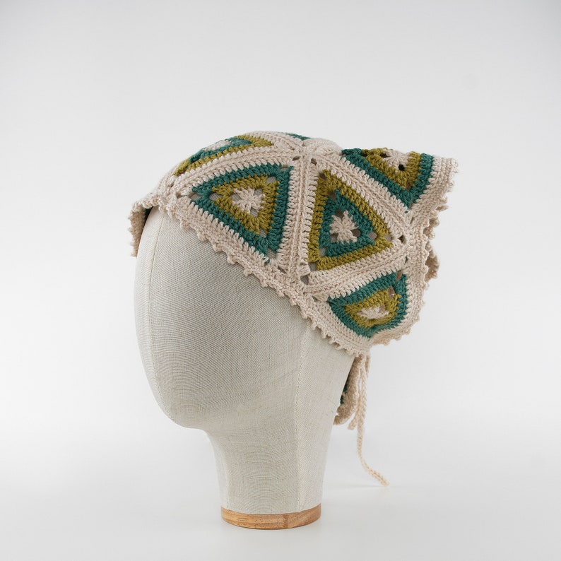 May include: A cream-colored crochet headscarf with a geometric pattern of green and olive triangles. The head covering is displayed on a mannequin head and has a scalloped edge and tie closures.