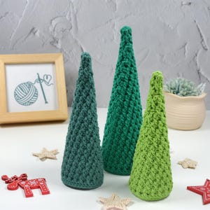May include: Three crocheted Christmas trees in different sizes and shades of green. The smallest tree is a light green, the middle tree is a darker green, and the largest tree is a teal green.